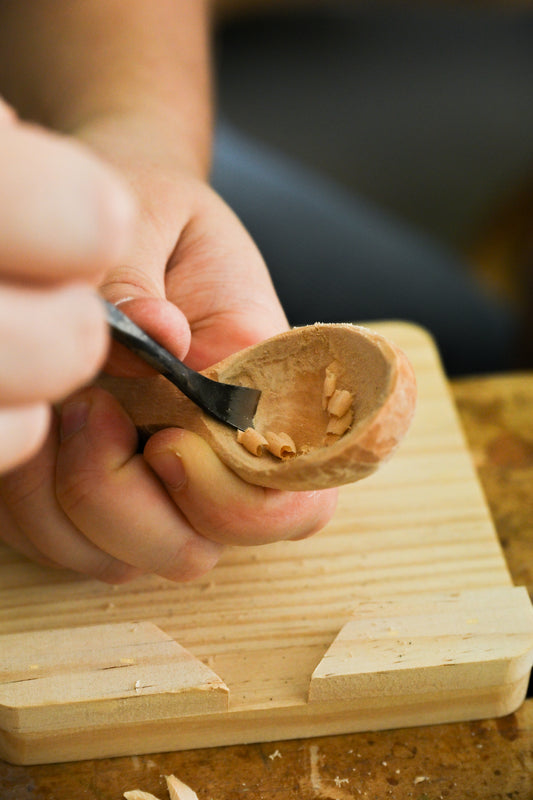 Hands using a carving knife to shape the bowl of a wooden spoon during the carving process.