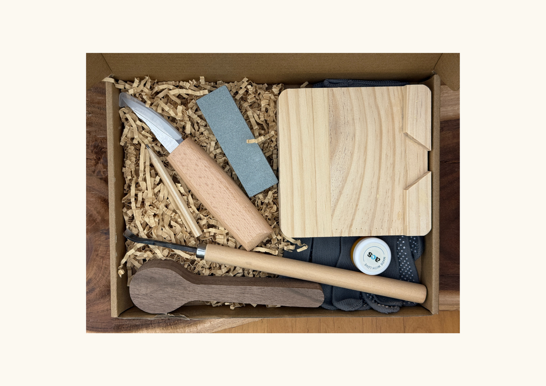 Spoon carving kit in a box with wooden blank, carving knives, sandpaper, gloves, and packing shreds.