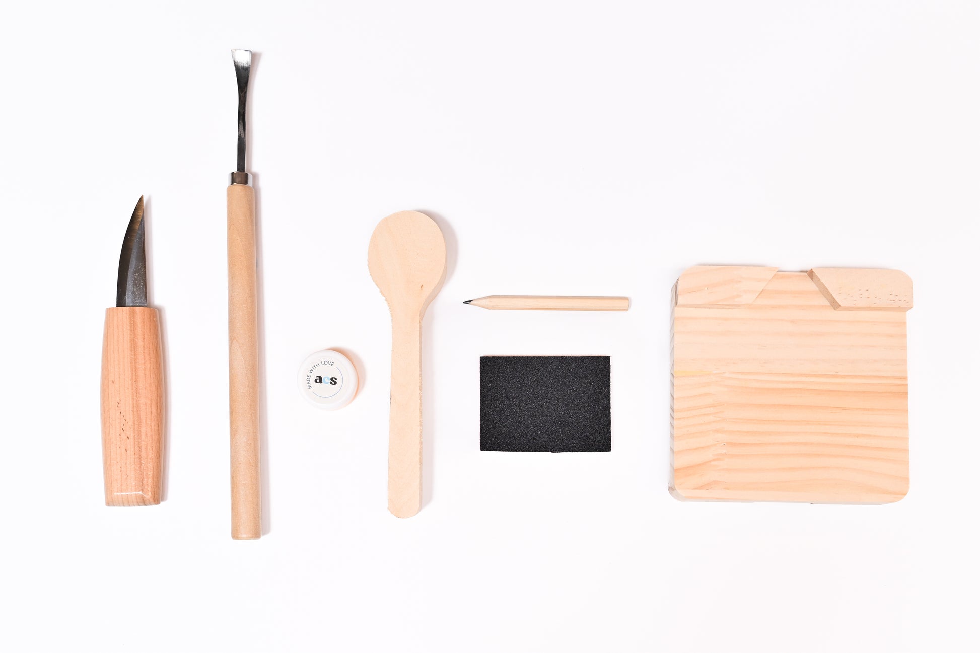 Beginner-friendly spoon carving kit with a wooden blank, carving tools, sandpaper and instructions arranged neatly on a white background.