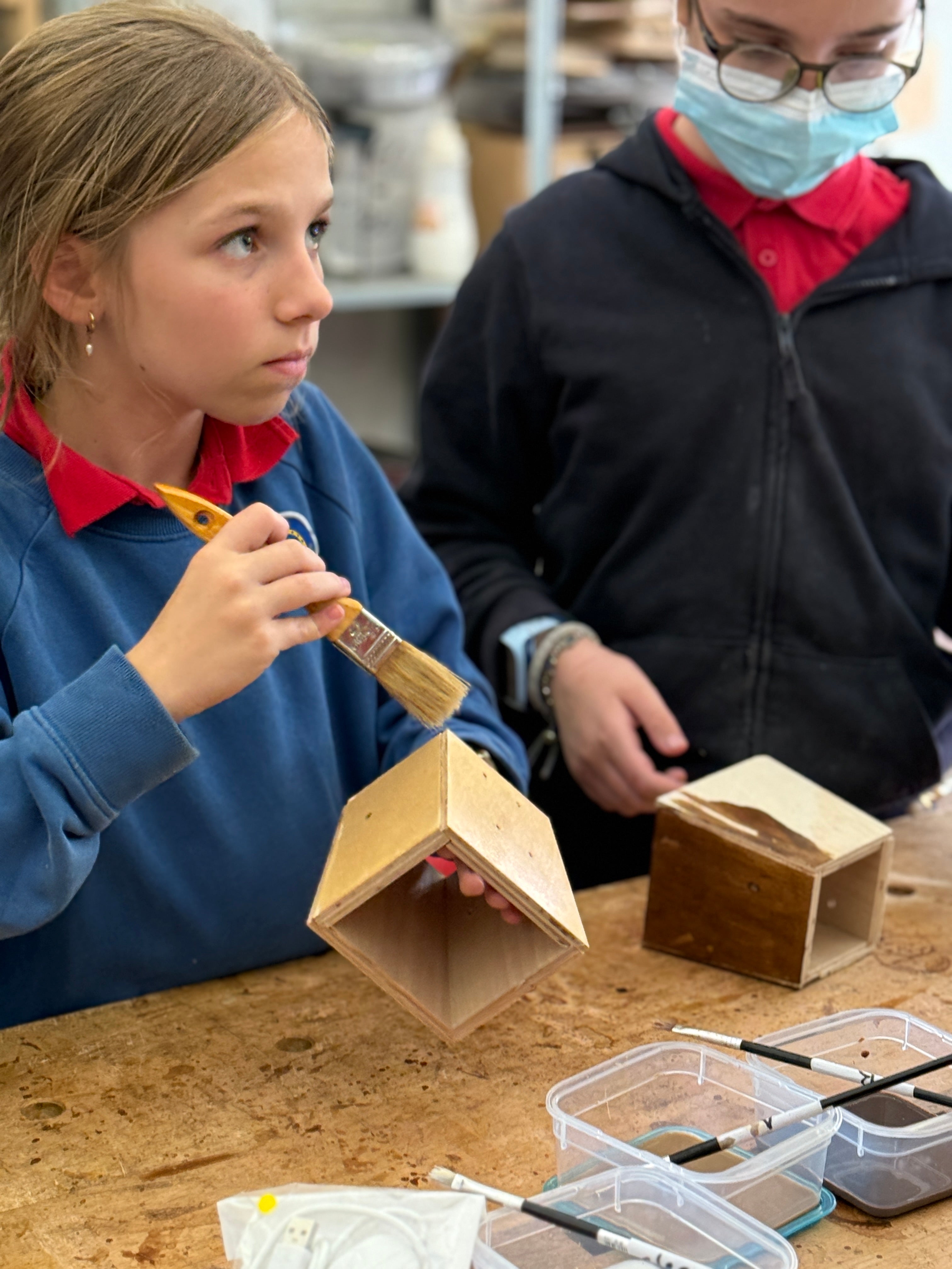 Two children working on a craft project with wooden boxes and paintbrushes in a classroom setting.
