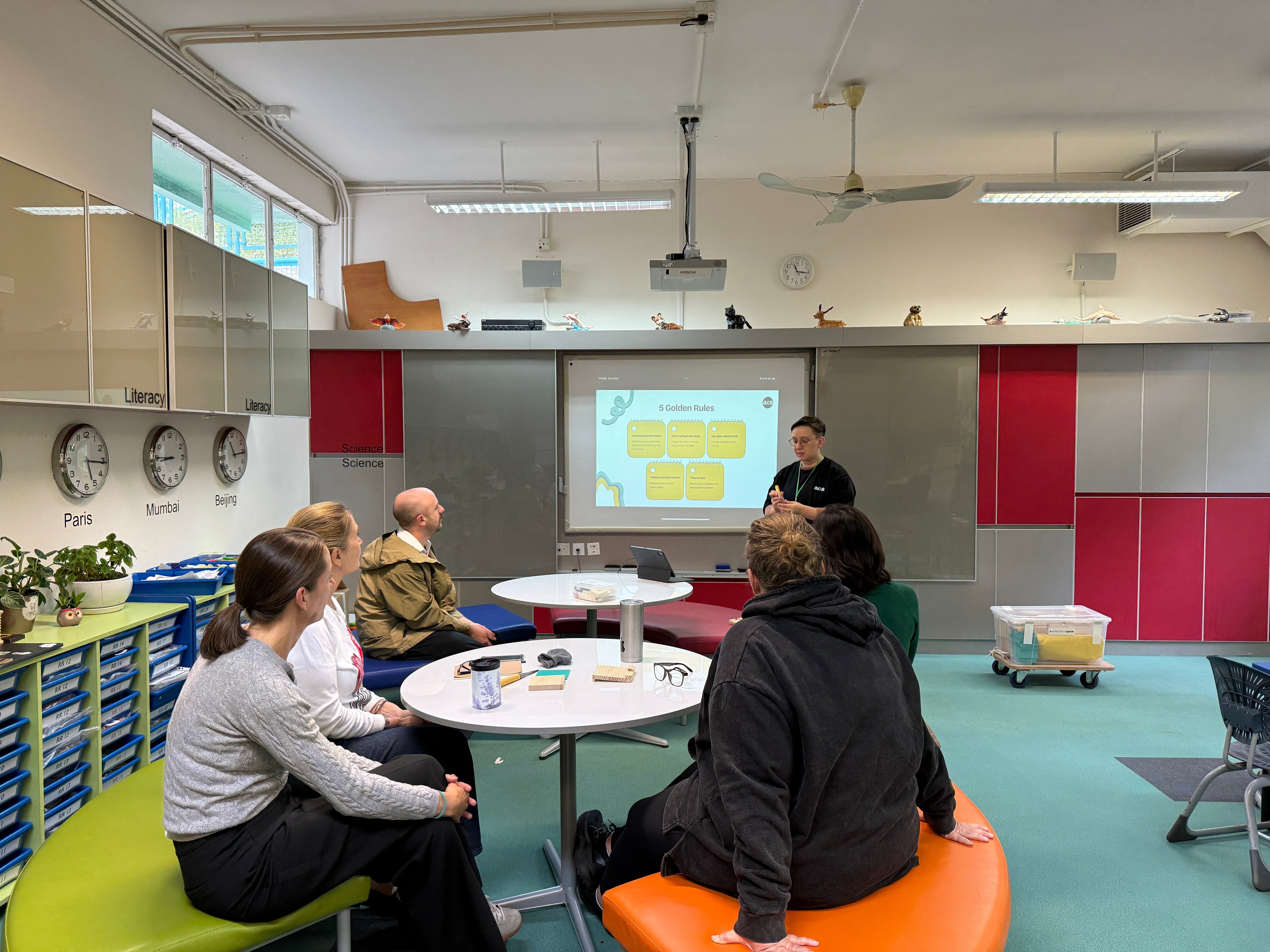 Group of people sitting on colorful chairs in a modern classroom setting with a presenter at the front.